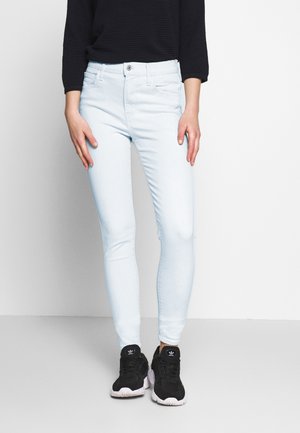 Personne porte un jean skinny bleu clair, un haut à manches longues noir et des baskets Adidas noires, debout devant un fond blanc.
