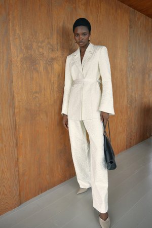 Costume texturé blanc avec un blazer ceinturé et un pantalon à jambes larges, associé à des escarpins beiges à bout pointu et un petit sac à main noir.