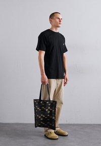 Geantă tote neagră cu modele galbene și maro, realizată din material texturat, cu două mânere rezistente. Persoană îmbrăcată cu un tricou negru și pantaloni bej.