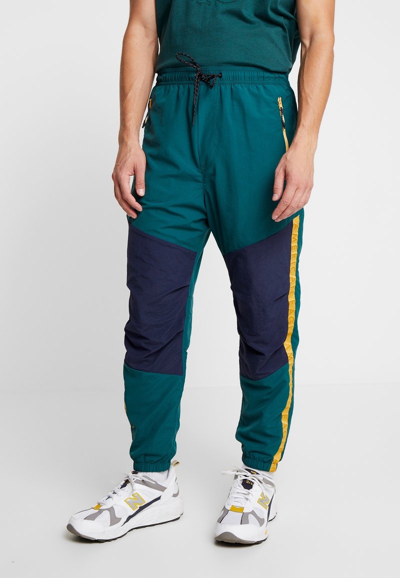 Homme portant un pantalon de jogging bleu canard avec des empiècements aux genoux bleu marine, des bandes latérales jaunes, une taille à cordon et des baskets blanches avec des touches de jaune.