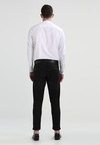 Bílá dlouhá rukávová košile s hladkým povrchem, doplněná obepínajícími černými džíny. Černé boty dokončují outfit. Zadní pohled.