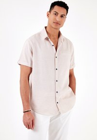 Camisa de botones de manga corta, de color beige claro y con tejido texturizado. Presenta un bolsillo en el pecho y botones en color marino, con los puños enrollados para un aspecto relajado.