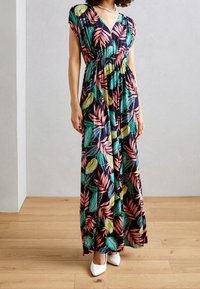 Kobieta w sukience bez rękawów o dekolcie w kształcie litery V, sięgającej do ziemi, w kolorowe tropikalne wzory liści, stojąca na drewnianej podłodze w białych czółenkach.