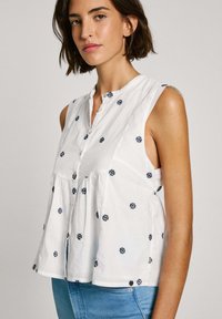 Blouse sans manches blanche avec broderie florale marine, fermeture à bouton et un design froncé à la taille pour un ajustement décontracté.