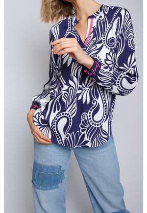 Femme portant un chemisier fleuri abstrait bleu marine et blanc avec des touches roses et un jean bleu clair avec un patch en denim effiloché sur la cuisse.