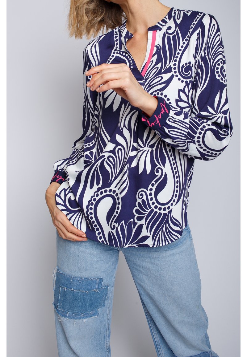 Femme portant un chemisier fleuri abstrait bleu marine et blanc avec des touches roses et un jean bleu clair avec un patch en denim effiloché sur la cuisse.