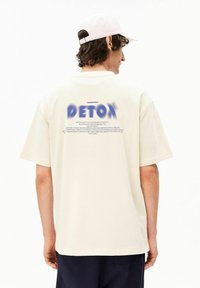 Roomwit oversized T-shirt met een blauwe verloopkleurige "DETOX"-logo op de achterkant, met tekst in een eenvoudig, schreefloos lettertype en korte mouwen.