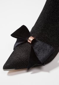 Chaussure à talons hauts noire avec un dessus pailleté, un bout pointu et un accent en velours texturé avec un nœud et une bande en rose doré.