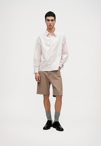 Camisa de botão listrada em rosa claro, com mangas longas, combinada com calções beiges até ao joelho. Sapatos pretos e meias cinzentas completam o traje.