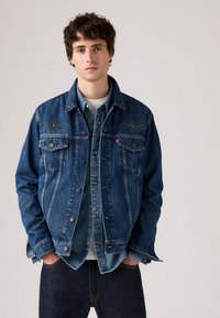 Donkerblauwe denimjacket met decoratieve stiksels en twee borstzakken, gelaagd over een lichter denimshirt met een wit onderhemd.