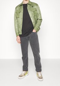 Mężczyzna w jasnoszarej pikowanej kurtce noszący czarną koszulę, ciemnoszare dżinsy i beżowe sneakersy z neonowożółtymi detalami, pozujący z jedną ręką w kieszeni.