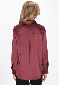 Cămașă din satin bordo, cu mâneci lungi, guler ascuțit și pliuri la spate. Dispune de manșete cu nasturi și tiv curbat pentru un croi lejer.