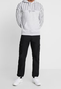 Felpa grigia con strisce verticali nere e tasca frontale, abbinata a pantaloni cargo neri con tasche laterali e sneakers bianche.