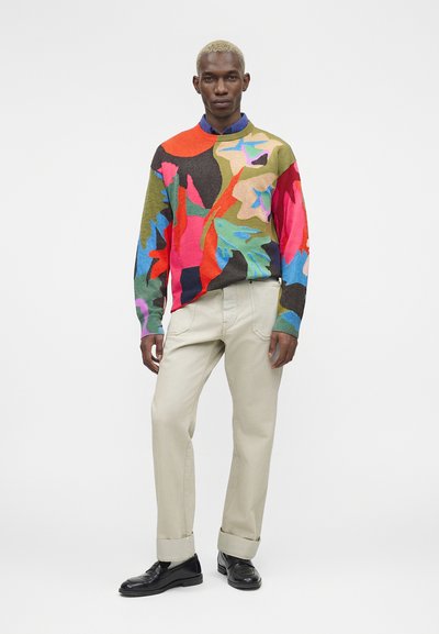 Άνδρας που φοράει πουλόβερ με πολύχρωμο αφηρημένο σχέδιο, μπεζ παντελόνι με μανσέτες, μαύρα μοκασίνια και μπλε πουκάμισο με γιακά από κάτω.