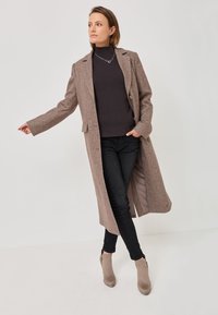 Manteau long en laine marron avec un col à revers, fermeture à un bouton, et poches latérales, porté sur un pull sombre et un jean noir.
