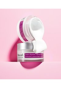 Die Murad Hydro-Dynamic Ultimate Moisture Creme in einem klaren Glasbehälter mit einem silbernen Deckel, der ein pinkes Etikett trägt, lässt weiße Creme auf einen pinken Hintergrund herauslaufen.
