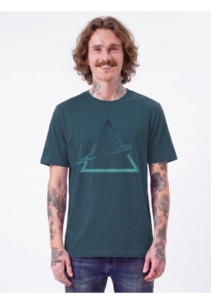 Mann mit schulterlangem Haar, Schnurrbart und Tätowierungen am Hals und an den Armen, der ein dunkelgrünes T-Shirt mit einem blau-grünen Dreiecksmuster trägt.