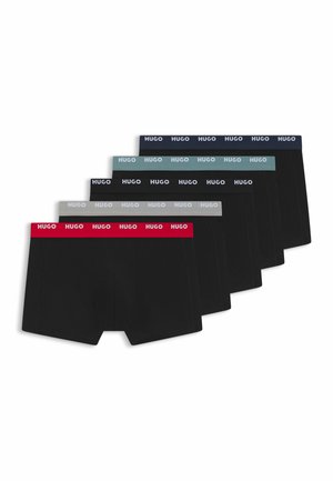 Jeune homme aux cheveux courts et foncés portant un boxer noir Hugo, debout devant un fond blanc uni.