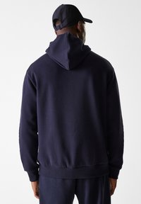 Dunkelblaues Kapuzensweatshirt mit weicher Textur. Es verfügt über eine Kängurutasche, gerippte Ärmelbündchen und Saum. Die Rückansicht zeigt ein großes Detail der Kapuze.