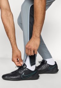 Grijze sportbroek met zwarte mesh-accenten en lichtblauwe bies; zwarte Nike schoenen met gestructureerd oppervlak en witte sokken. De broek aanpassen.