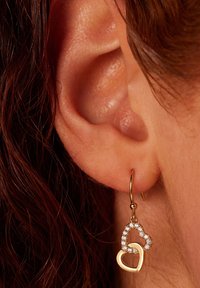 Des boucles d'oreilles en forme de cœur en or présentent un double design, l'une étant contourée et l'autre ornée de pierres transparentes scintillantes, suspendue à un crochet.