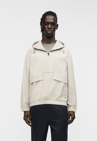 Μπεζ ελαφρύ μπλουζάκι με κουκούλα, εμπρόσθιο φερμουάρ και δύο μεγάλες μπροστινές τσέπες. Διαθέτει ελαστικό τελείωμα και λείο υφαντό υλικό.