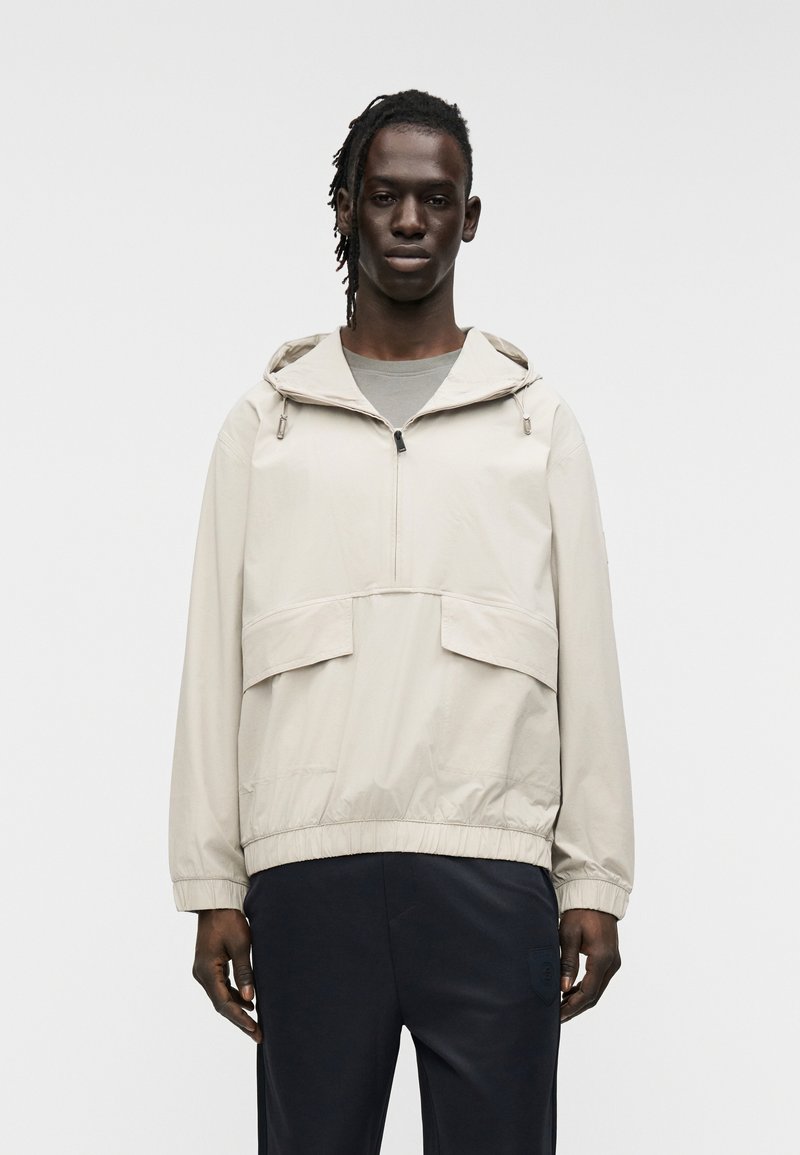 Μπεζ ελαφρύ μπλουζάκι με κουκούλα, εμπρόσθιο φερμουάρ και δύο μεγάλες μπροστινές τσέπες. Διαθέτει ελαστικό τελείωμα και λείο υφαντό υλικό.