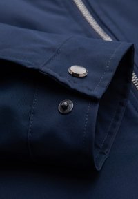 Gros plan d'un poignet de veste bleu marine avec un bouton-pression argenté et un fermoir pression noir sur un tissu lisse.