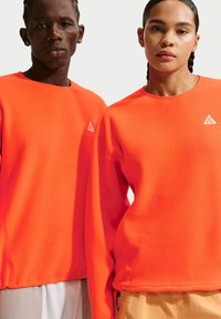 Δύο μοντέλα φορούν φωτεινά πορτοκαλί μακρυμάνικα μπλουζάκια με μικρά λευκά τριγωνικά λογότυπα στο στήθος, μπροστά σε απλό φόντο.