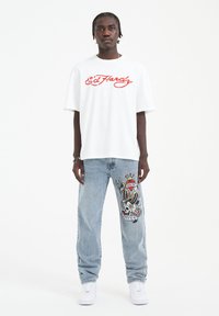 T-shirt oversize bianco con logo rosso "Ed Hardy", abbinato a jeans in denim azzurro chiaro con design ricamato di teschio e serpente colorato.