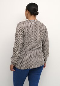 Langærmet top i lys brun med et geometrisk hvidt mønster, der har en tætsiddende talje og let sammensnørede ærmer. Matchende med blå jeans.