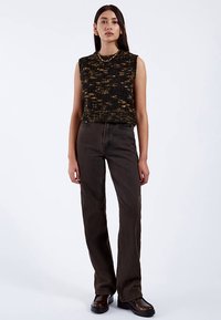 Schwarzes und goldenes, strukturiertes ärmelloses Oberteil, kombiniert mit braunen Schlagjeans und schwarzen Plateauschuhen. Schlichte Accessoires runden das Outfit ab.