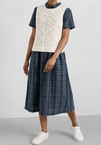 Kremowa dzianinowa kamizelka z geometrycznymi wzorami, nałożona na ciemnoniebieską sukienkę w kratę. Białe trampki dopełniają strój.