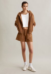 Sweat à capuche zippé en éponge brun associé à un short assorti. Top court côtelé blanc en dessous. Le mannequin porte des baskets blanches et des chaussettes de sport.