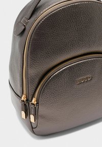Dunkler Leder-Rucksack mit doppelten goldenen Reißverschlüssen und runder Form. Logo "LIUJO" auf der Vorderseite geprägt. Strukturierte Oberfläche sichtbar.