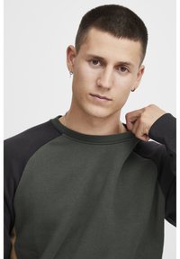 Mørkegrøn sweatshirt med sorte raglanærmer og ribbet krave. Glat tekstur, har en rund halsudskæring og diskrete stiklinger.