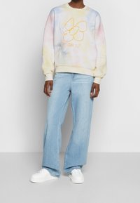 Persoon die een pastel tie-dye sweatshirt draagt met een gele bloem en tekst, lichtblauwe wijde jeans en witte sneakers tegen een effen achtergrond.