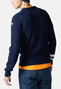 Marinblå stickad tröja med rund halsringning, ribbade ärmmuddar och vita randaccent på ärmarna. Bärs över en orange t-shirt och blå byxor.