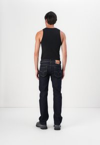 Jeans de mezclilla azul oscuro con un diseño de pierna recta, usados con una camiseta sin mangas negra. Presenta un parche de cuero en la cinturilla.