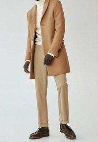 En tan uldfrakke over en cremet turtleneck, parret med beige bukser og brune snørebåndssko. Har en skræddersyet pasform og struktureret silhuet.