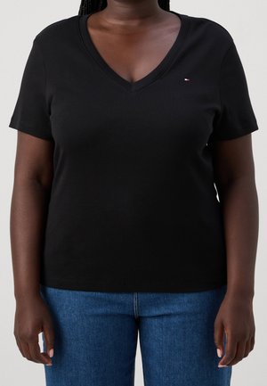 Persoon in een zwart V-hals shirt met korte mouwen en blauwe jeans, staand tegen een neutrale achtergrond.