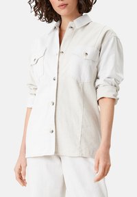 Camisa ligera de botones en tonos beige y blanco. Presenta un cuello clásico, dos bolsillos en el pecho y mangas enrolladas. Textura suave.
