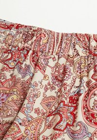 Gros plan sur un tissu avec une ceinture élastique présentant un motif détaillé de paisley et floral en rose, rouge, violet et jaune sur fond crème.