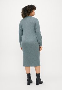 Stickad klänning i mjuk teal med ribbad textur, lång ärm och en figurnära siluett som slutar nedanför knäet. Matchas med svarta stövlar med blockklack.