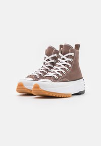 Brązowe sneakersy za kostkę wykonane z płótna, z białymi sznurówkami i kontrastową białą podeszwą. Teksturowana gumowa podeszwa zewnętrzna ma zygzakowaty wzór dla lepszej przyczepności.