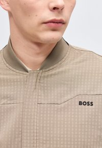 Νέος άνδρας που φοράει ένα μπεζ καρό σακάκι με ριμπ γιακά και λογότυπο "BOSS" στο στήθος, εμφανιζόμενος από το λαιμό έως το πάνω μέρος του στήθους.