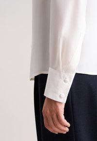 Blusa bianca a maniche lunghe con una texture liscia, dotata di polsini con bottoni e una vestibilità leggermente ampia, abbinata a pantaloni scuri.