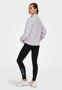 Pullover lilla con colletto alto, abbinato a leggings neri con testo bianco. Il modello indossa sneakers bianche.