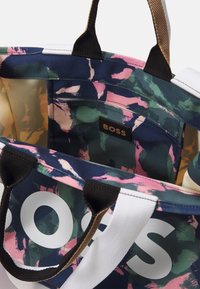 Bolso tote de lienzo con un patrón abstracto en verde, rosa y azul marino, texto en blanco "BOSS" en el exterior, y bolsillo interior con cremallera y etiqueta "BOSS".
