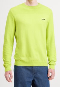 Man som bär en limegrön långärmad stickad tröja med ribbade ärmar och en svart "BOSS"-logotyp på bröstet, tillsammans med mörkblå byxor.